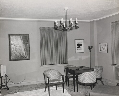 A black-and-white photograph of a corner seating area in a hotel room showing three paintings hanging on the wall between windows.