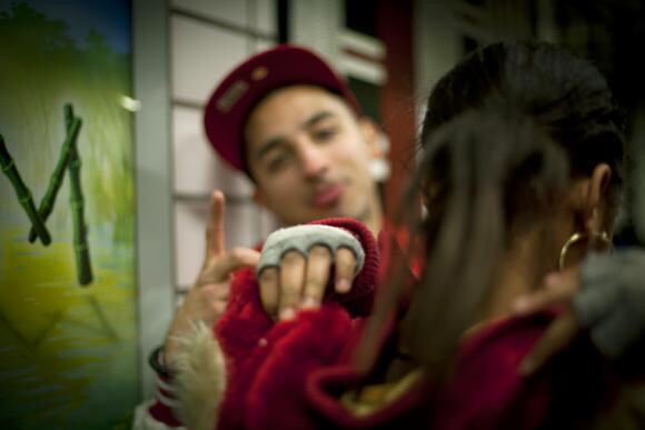 A color photograph of two young people: one from the back getting ready to pull a hood over their head, the other wearing an upturned baseball hat facing the first person