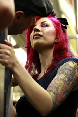 A color photograph of a light-skinned woman with pink hair and tattoos on her arm holds a stanchion on a crowded subway train. 