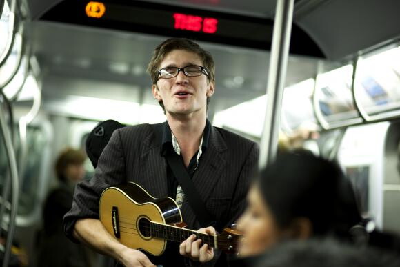 A color photograph of a White man with glasses playing a ukulele and singing inside of a subway train.