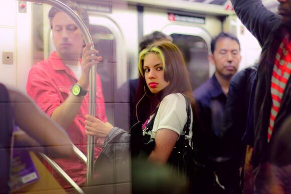 A color photograph of people standing in a subway train, most holding handrails above and in front of them.