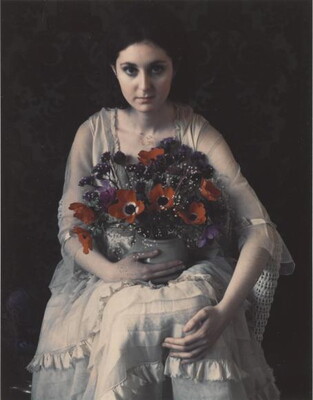 A color photograph of a seated White woman with dark hair wearing a white dress holding a vase of poppies in her lap.