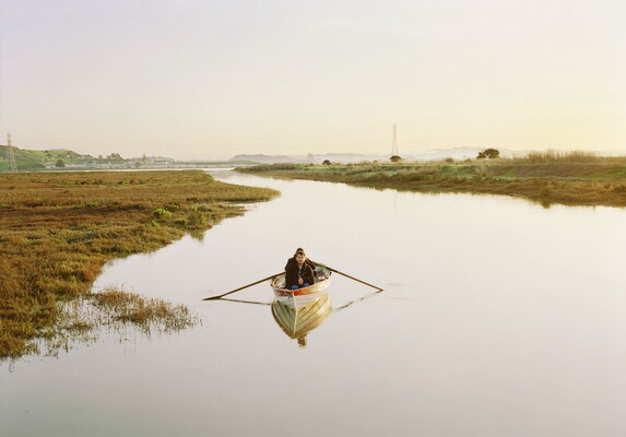 A color photograph of two White people in a rowboat on a still river with a small town in the distance.
