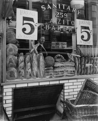 A black-and-white photograph of a bakery shop window with loaves of bread stacked in displays and signs that read, "Bread 5 cents a loaf."