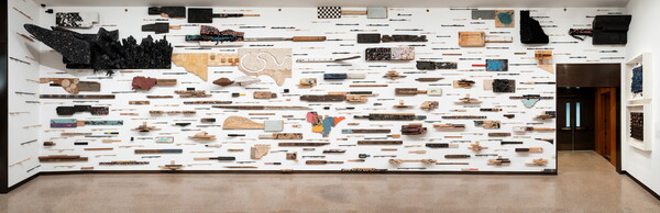 An installation view of a gallery wall covered in a grid of relief sculptures of wood, paper, metal, and textiles with varied color and texture.