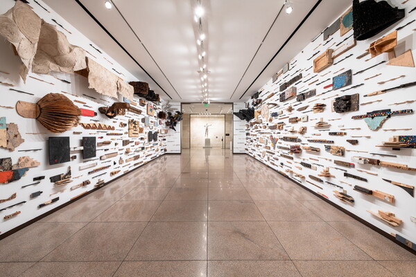 An installation view of a gallery wall covered in a grid of relief sculptures of wood, paper, metal, and textiles with varied color and texture.