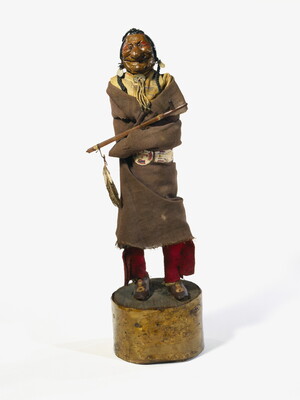 A painted wood sculpture of a smiling Native American man with exaggerated features wrapped in a blanket with a pipe resting on his arm.