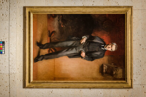 A life-size oil painting of an middle-aged man in a dark, three-piece suit and tie with his thumbs in his front pockets, standing in front of a large fireplace.