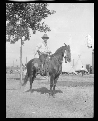 A black-and-white image of a man wearing a cowboy hat seated on a horse; several tipis in the background.