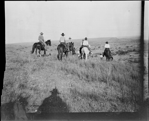 A black-and-white image of a group of cowboys on horseback walking away from the viewer toward a field.