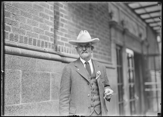 A black-and-white image of an older White man wearing a cowboy hat and a three-piece suit, holding a cigar as he stands in front of a brick building.
