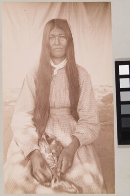 A sepia-toned portrait of a seated Indigenous woman with long dark hair wearing a long-sleeved gingham dress.