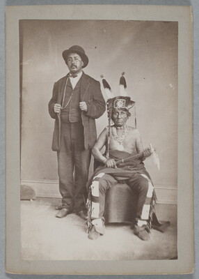 A black-and-white photograph of two Indigenous men: the one standing is wearing a bowler hat and a three-piece suit; the one seated wears a feathered hat, many necklaces, fringed pants, and holds a tomahawk in his lap.