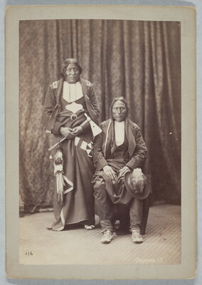 A sepia-toned portrait photograph of two Indigenous people: one seated with a hat on their knee, and the other standing holding a tomahawk.