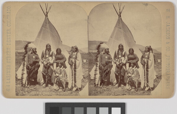 A black-and-white stereograph of five Indigenous people in traditional regalia standing in front of a tipi.
