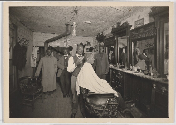 A black-and-white snapshot of the inside of a barber shop with a Black man in a chair getting his hair cut.