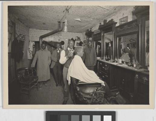 A black-and-white snapshot of the inside of a barber shop with a Black man in a chair getting his hair cut.