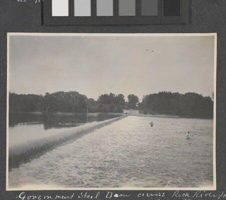 A black-and-white photograph of people fishing in a rushing river near a dam.