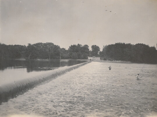A black-and-white photograph of people fishing in a rushing river near a dam.