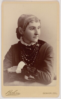 A black-and-white portrait photograph of a young White woman, hair pulled back and short bangs framing her face, wearing a dark dress with a ruffled white collar and sleeves, leaning with her elbows on the surface in front of her.