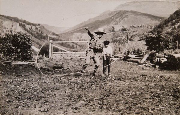 A black-and-white photograph of a White cowboy spinning a lasso for a child in a rural fenced area.
