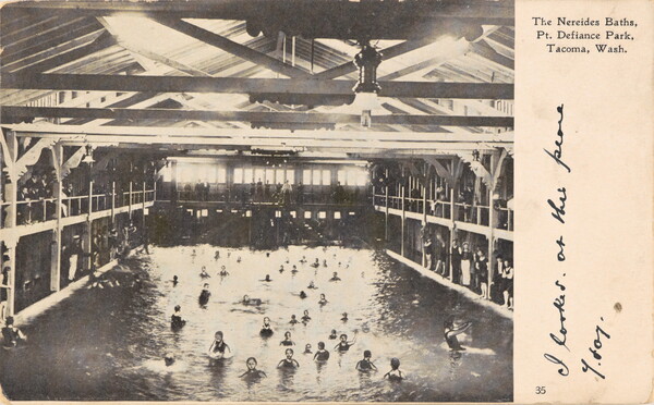 A black-and-white postcard of people in a large swimming pool inside a high-roofed, multi-level building.
