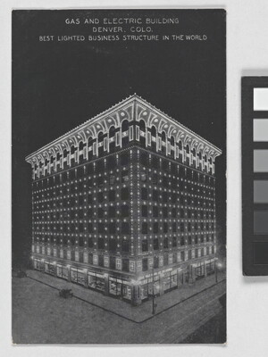 A color postcard of a nine-story building lit up at night under a full moon that is partially hidden behind clouds.