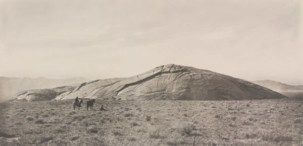A black-and-white photograph of two people and two horses in a grassy field in front of a very large, smooth rock that seems to be emerging from the ground.