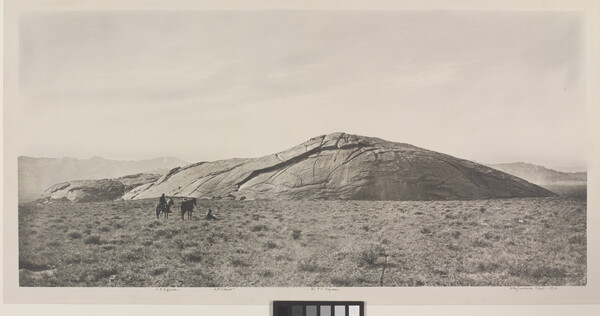 A black-and-white photograph of two people and two horses in a grassy field in front of a very large, smooth rock that seems to be emerging from the ground.