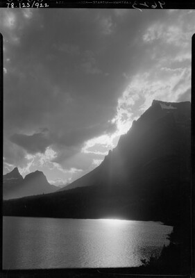 A black-and-white image of a cloudy sky with the sun setting behind a mountain overlooking a still lake.