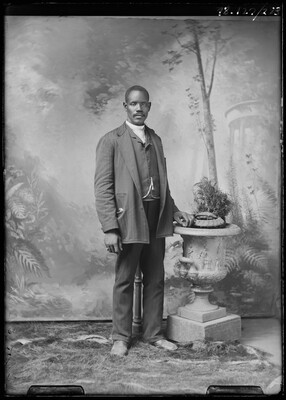 A black-and-white studio image of a standing Black man with a mustache, wearing a suit and vest, a watch chain dangling at his waist, next to a large urn.
