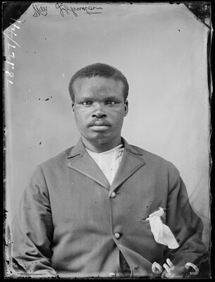 A black-and-white studio image of a seated young Black man wearing a suit jacket, a white pocket square stuffed into a breast pocket, over a white shirt.