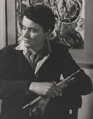 A black-and-white portrait photograph of a seated White woman holding paintbrushes in front of an abstract painting.