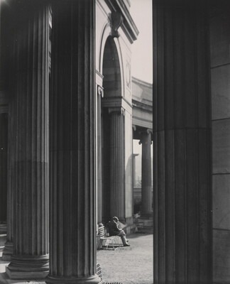 A black-and-white photograph of a person sitting on a bench reading a newspaper as seen from between large fluted columns.