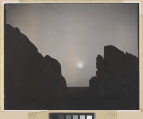 A black-and-white photograph of a moon shining through a foggy sky from between the V-shaped silhouette of large rocks.