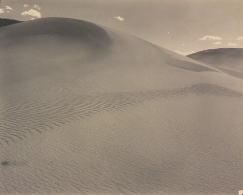 A sepia-toned photograph of a rippling sand dune rising up into a sky dotted with a few small clouds.