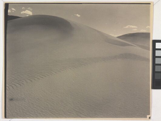A sepia-toned photograph of a rippling sand dune rising up into a sky dotted with a few small clouds.