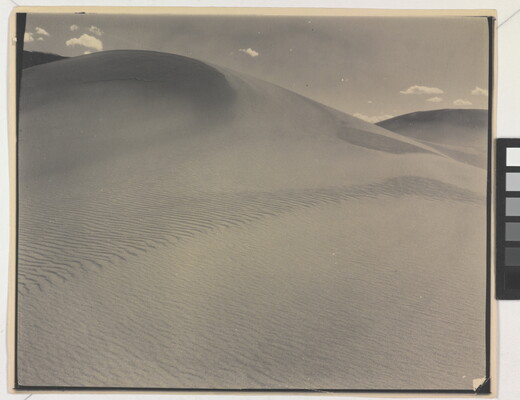 A sepia-toned photograph of a rippling sand dune rising up into a sky dotted with a few small clouds.