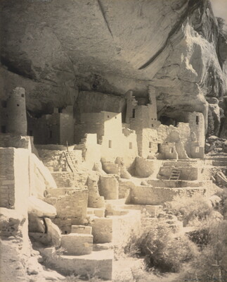 A black-and-white photograph of an abandoned, crumbling rock city built into the side of a desert cliff.