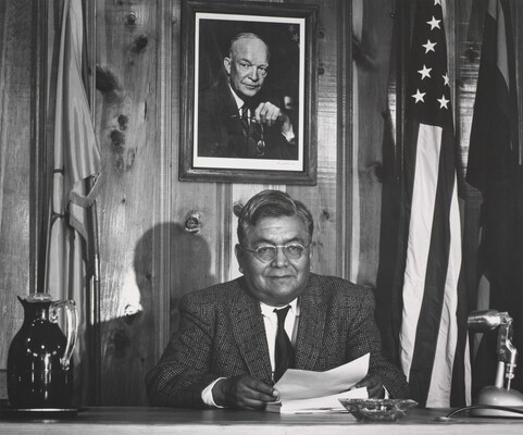 A black-and-white photograph of an Indigenous man dressed in a suit, seated at a desk between two flags and under a portrait of President Eisenhower.