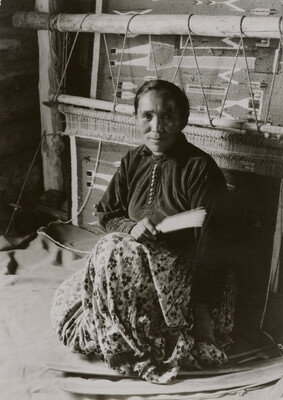 A black-and-white photographic portrait of a Native American woman sitting on the floor in front of a large loom.