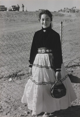A black-and-white photograph of a young Indigenous woman wearing a long white skirt and dark blouse and standing in front of a chain link fence.
