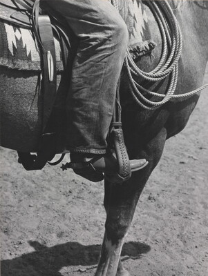 A black-and-white photograph of a person's leg in a cowboy boot, including spurs, sitting astride a horse, viewing only the horse's front quarter.