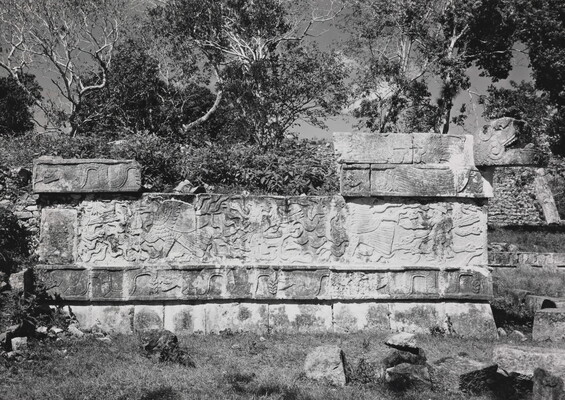 A black-and-white photograph of a crumbling stone wall engraved with designs surrounded by grass and trees.