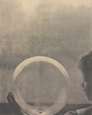 A black-and-white photograph of a White child looking at a clear glass globe in front of a rain-spattered window.