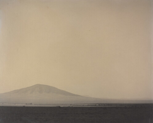 A black-and-white photograph of a flat, desert landscape with a mountain rising up in the hazy distance.