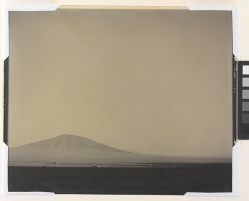 A black-and-white photograph of a flat, desert landscape with a mountain rising up in the hazy distance.