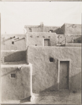 A black-and-white photograph of a multi-level adobe pueblo with wood doors in some of the walls.
