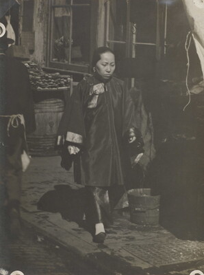 A black-and-white photograph of an woman with Asian features wearing a billowing tunic over pants walking down a commercial street.