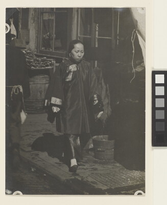 A black-and-white photograph of an woman with Asian features wearing a billowing tunic over pants walking down a commercial street.